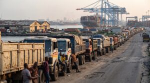 Trucks queue outside a Nigerian port with stacked containers in the background.